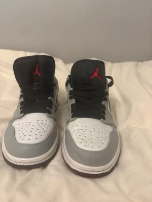 Air Jordan 1 Low Vit/Grå/Svart - Säljer ett par Air Jordan 1 Low i vitt, grått och svart med klassisk röd Jumpman-logga på plösen. Skorna har svart swoosh, svarta skosnören och perforerad tåbox. Ovandelen är i läder och designen är riktigt clean och tidlös.