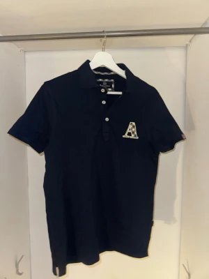 aquascutum pike marinblå - Snygg marinblå pikétröja från Aeronautica Militare med korta ärmar, krage och knappar framtill. Stor patch med bokstaven A på bröstet och en brittisk flagga på ena ärmen. Tillverkad i mjuk bomull för skön känsla.