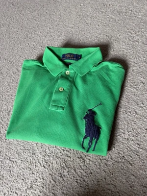 Grön piké från Polo Ralph Lauren - Snygg grön pikétröja från Polo Ralph Lauren med klassisk krage, två knappar och stor mörkblå logga broderad på bröstet. På vänster ärm finns en stor mörkblå siffra 3. Perfekt till sommaren och enkel att styla.