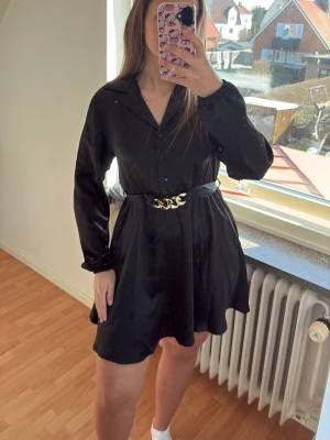 Svart kort klänning med bälte - Svart kort klänning med lång ärm och krage, knappar framtill och ett snyggt bälte med gulddetalj i midjan. Klänningen har en lätt utsvängd form och passar perfekt för dig som gillar stilrena och trendiga plagg. Felfri!