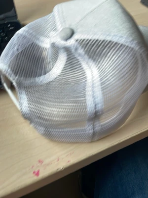 Vit truckerkeps med tryck framtill - Snygg vit truckerkeps med mesh på sidorna och baksidan för extra ventilation. Framtill finns ett svartvitt tryck med motiv. Justerbar storlek baktill med plastspänne. Perfekt för en avslappnad streetstyle-look.