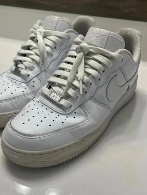 Nike Air Force 1 vita sneakers - Klassiska Nike Air Force 1 sneakers i helvitt läder med perforerad tåbox, vit sula och ikonisk Swoosh-logga på sidorna. Skorna har platta vita skosnören och låg siluett som ger en clean och tidlös look.