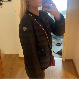 Svart pufferjacka från Moncler - Snygg svart pufferjacka från Moncler med quiltad design och dragkedja framtill. Jackan har två fickor med dragkedja och Moncler-logga på ärmen. Perfekt för våren oxh när de är lite kallare