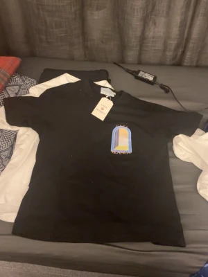 Svart t-shirt från Casablanca - Svart t-shirt från Casablanca med färgglatt tryck på bröstet och stor grafisk design på ryggen. T-shirten har rund hals och korta ärmar. Perfekt för dig som gillar streetwear med unik touch.