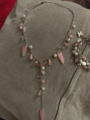 Halsband&armband med rosa och vita pärlor - Charmigt armband med mix av rosa och vita pärlor i olika storlekar och former. Armbandet har detaljer i metall och små blommor som ger en söt och lekfull känsla. Perfekt accessoar för dig som gillar romantiska och feminina smycken.