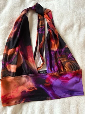Halterneck top från NLY One - Säljer en snygg halterneck top från NLY One med färgstarkt abstrakt mönster i lila, orange och svart. Modellen har halterneck med knyt i nacken och bred underbystresår för extra stöd i storlek M