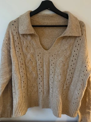 Beige kabelstickad tröja med krage - Säljer en beige stickad tröja med bred krage och v-ringning. Tröjan har kabelstickat mönster framtill och dekorativa hålstickade detaljer. Ribbstickade muddar vid ärmslut och nederkant. Perfekt för dig som gillar en avslappnad men ändå snygg stil.