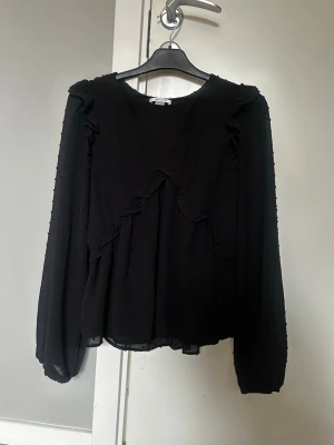 Svart blus med volanger från Gina Tricot - Snygg svart blus från Gina Tricot med volangdetaljer framtill och diskret prickstruktur i tyget. Blusen har lång ärm och är tillverkad i ett tunt, lätt material som ger ett luftigt intryck. Perfekt att styla med jeans eller kjol.