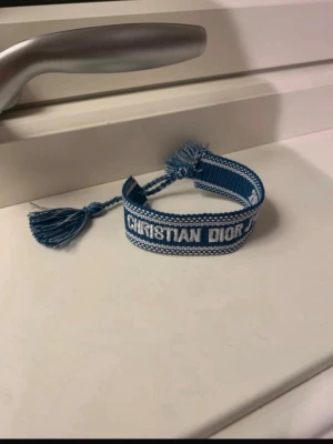 Blått armband från Christian Dior - Snyggt blått armband från Christian Dior med vit brodyr och tofsar i ändarna. Armbandet är vävt i tyg och har märkets logga tydligt broderad. Perfekt accessoar för att lyfta din outfit med en exklusiv touch.