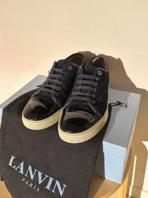 Svarta Lanvin sneakers med lackdetalj - Snygga svarta sneakers från Lanvin med ovandel i mocka och glansig lackad tå. Skorna har vita sulor och svarta skosnören. Klassisk låg modell med stilren design och diskreta metalldetaljer. Perfekta för dig som gillar exklusiva och trendiga sneakers.