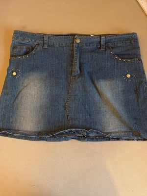 Vintage jeanskjol med nitar - Snygg blå jeanskjol med kort längd och coola nitar vid fickorna. Kjolen har klassisk femficksdesign, knapp och dragkedja framtill. Perfekt för dig som gillar en avslappnad och trendig stil. 