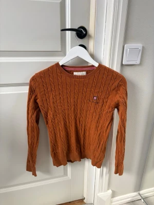 Rustbrun kabelstickad tröja Bondelid - Säljer en rustbrun kabelstickad tröja från Bondelid. Tröjan har rund halsringning, långa ärmar och ett litet broderat märke på bröstet. Klassisk och tidlös design med ribbade muddar och nederkant. Perfekt för lager-på-lager-stil.