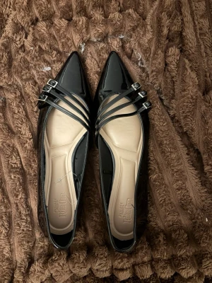 Svarta lackade ballerinaskor med remmar - Snygga svarta ballerinaskor i lack med spetsig tå och tre tunna remmar med silverspännen över foten. Skorna har en stilren och modern design med beige innersula för extra komfort. Perfekta för dig som gillar en trendig och elegant look.