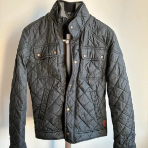 Quiltad svart jacka från Jack&Jones - Snygg svart quiltad jacka från Vintage med guldiga knappar och hög krage. Jackan har två bröstfickor med lock och tryckknappar samt markerade sömmar i rutmönster. Perfekt för dig som gillar stilren och tidlös design.