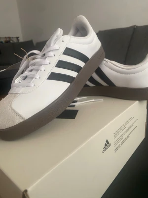 Adidas vita sneakers med bruna sulor - Snygga vita Adidas sneakers med klassiska svarta ränder på sidorna och brun gummisula. Ovandelen är i läder och mocka vid tån, med vita skosnören och diskret Adidas-logga på hälen. Perfekta för en clean och sportig look.