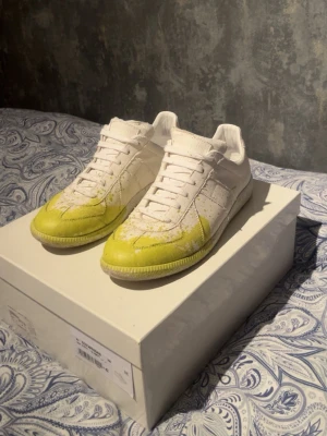 Maison Margiela gula och vita sneakers - Unika sneakers från Maison Margiela i vitt läder med distinkt gul färg på tån och sulan. Klassisk låg modell med vita skosnören och subtila detaljer. Perfekta för dig som vill sticka ut med en kreativ och trendig look.