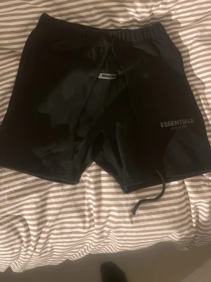 Svarta Essentials shorts   - Säl reaching ett par svarta shorts från Essentials med snörning i midjan och diskret logga på vänster ben. Shortsen har elastisk midja och en bakficka. Perfekta för chill dagar eller trä allmänna aktiviteter. Vid märket i mitten finns det en liten svart märke där!