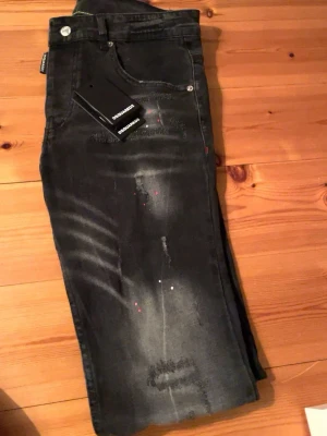 Svarta Dsquared2 jeans med detaljer - Säljer ett par svarta jeans från Dsquared2 med coola slitningar och färgstänk. Jeansen har klassisk femficksmodell, silverfärgade knappar och en röd logotyppatch bak i midjan. Materialet är denim med stretch för skön passform. Perfekta för dig som gillar streetstyle.