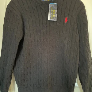 Svart kabelstickad tröja Polo Ralph Lauren - Kabelstickad svart tröja från Polo Ralph Lauren med röd broderad logga på bröstet. Tröjan har rund halsringning och ribbade muddar vid ärmslut och nederkant. Klassisk och stilren design som passar perfekt till många outfits.