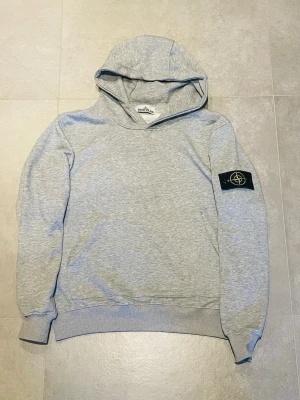 Stone Island hoodie - Snygg ljusgrå hoodie från Stone Island med klassisk logotyp-patch på vänster ärm. Hoodien har huva, ribbade muddar och är tillverkad i mjukt material som passar perfekt till en avslappnad stil.den är  f ake 