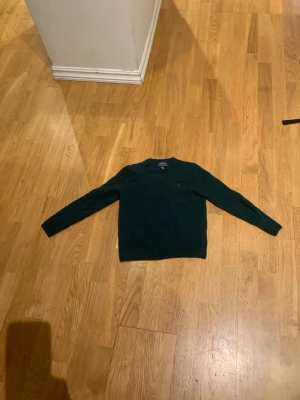 Mörkgrön stickad tröja Polo Ralph Lauren - Mörkgrön stickad tröja från Polo Ralph Lauren med rund halsringning och långa ärmar. Klassisk och enkel design utan mönster, perfekt för lager på lager. Tröjan har en diskret logga på bröstet och är gjord i mjukt material.