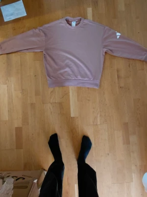 Rosa sweatshirt från Adidas - En baggy rosa sweatshirt från Adidas med rund hals och långa ärmar. Tröjan har en enkel design med en liten vit Adidas-logga på ärmen. Perfekt för en avslappnad och trendig look.