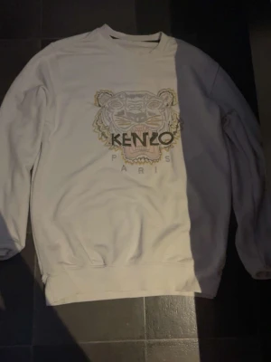 Vit sweatshirt från Kenzo med tigertryck - Säljer en vit sweatshirt från Kenzo med det ikoniska broderade tigerhuvudet och texten 'KENZO PARIS' på bröstet. Tröjan har rund halsringning, långa ärmar och ribbade muddar. Perfekt för dig som gillar streetwear och snygga loggor.