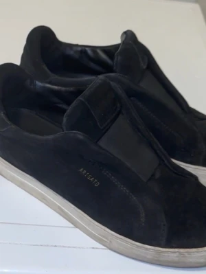 Svarta sneakers från Axel Arigato - Stilrena svarta sneakers från Axel Arigato i mocka med elastiska paneler istället för snörning. Skorna har en låg profil och vit sula, samt diskret logga i guld på sidan. Perfekta för en clean och modern look.