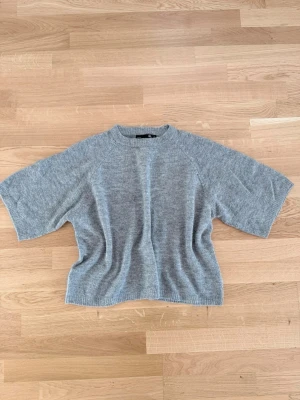 Grå stickad tröja - Mysig grå stickad tröja med korta, vida ärmar och rund hals. Tröjan har en avslappnad passform och ribbade kanter vid ärmslut och nederkant. Perfekt för lager på lager och enkel att matcha med olika stilar.