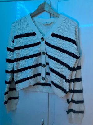 Vit och svart randig kofta med knappar - Snygg stickad kofta i vitt med svarta horisontella ränder och stora knappar framtill. Koftan har v-ringning, långa ärmar och ribbade muddar. Perfekt för lager-på-lager och en chill look.