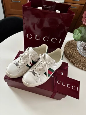 Gucci vita sneakers med gröna/röda ränder - Snygga vita sneakers från Gucci med klassiska gröna och röda ränder på sidorna. Skorna har vita snören, metall-detaljer framtill och grön detalj på hälen. Tillverkade i läder och har en tidlös, clean design som passar till många outfits.