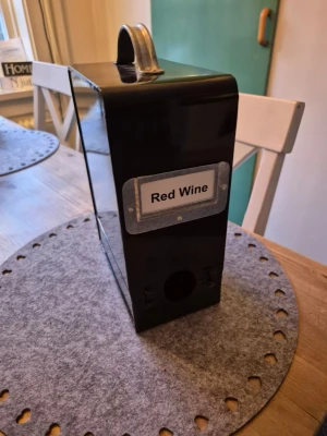 Svart boxvinslåda i metall - Snygg svart boxvinslåda i metall med handtag upptill och etikett där det står 'Red Wine'. Perfekt för att dölja och servera boxvin på ett stilrent sätt. Lådan har en öppning för kran och passar bra på bordet vid middagar.