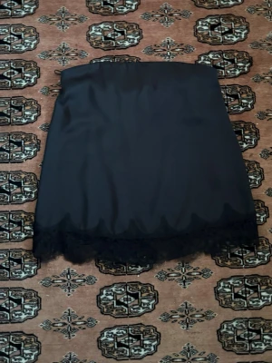 Svart bandeau topp med spetskant - Säljer en svart axelbandslös bandeau topp med snygg spetskant nedtill. Toppen har en slät, glansig yta och sitter löst över kroppen. Från Zara. Helt oanvänd då jag köpte i större storlek som satt bättre. 