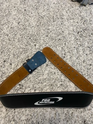 Brunt träningsbälte Pro Nutrition - Snyggt och robust träningsbälte från Pro Nutrition i brunt skinn med svart utsida och vit kontrastsöm. Perfekt för dig som vill ha extra stöd vid gymmet. Justerbar storlek med flera hål och metallspänne.