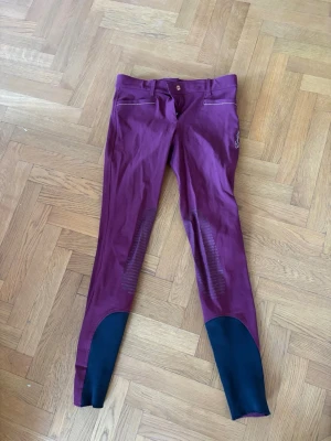 Vinröda equiline ridbyxor med svarta detaljer - Snygga vinröda equiline ridbyxor med svarta förstärkningar vid bensluten och silikongrepp på insidan av benen. Byxorna har dragkedjor fram och bak samt bälteshällor. Perfekta för dig som vill ha stil och funktion i stallet. Storlek IT 42. 