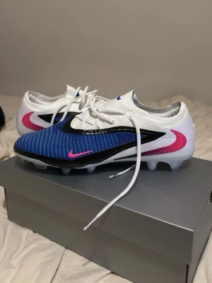 Nike Mercurial blå/vita fotbollsskor 46  - Snygga Nike Mercurial fotbollsskor med blå ovandel, vita sidor och rosa detaljer. Skorna har svart swoosh och är designade för spel på gräs. Lätta och smidiga med snörning och greppvänlig sula. Perfekta för dig som vill sticka ut på planen. Helt nya!