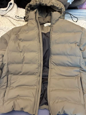 Beige pufferjacka från Pondus - Snygg beige pufferjacka från Pondus med huva och dragsko. Jackan har en quiltad design, dragkedja framtill och två sidofickor. Perfekt för kalla dagar när du vill hålla dig varm och stilren. Materialet är syntetiskt och jackan har en klassisk, boxig passform. Använd en gång endast. 