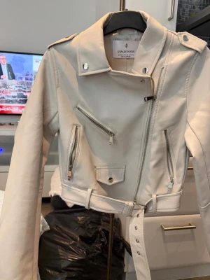 Vit skinnjacka från Stradivarius - Snygg vit skinnjacka från Stradivarius med klassisk bikerstil. Jackan har asymmetrisk dragkedja, flera silverfärgade dragkedjor och fickor samt ett bälte i midjan. Kortare modell som ger en trendig look och passar perfekt till jeans eller klänning.