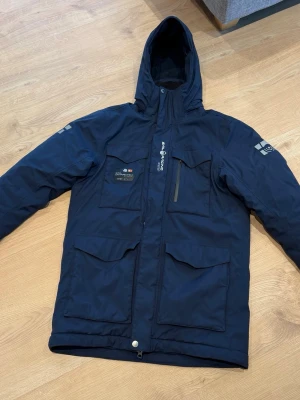 Sail racing vinterjacka  - Snygg mörkblå parkas från Sail racing med huva och flera praktiska fickor framtill. Jackan har dragkedja, tryckknappar och logga på ärmen. Materialet är tåligt och vattenavvisande, perfekt för kalla dagar. Stilsäker och funktionell design. Ny pris 4500 kr