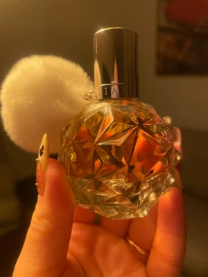Ariana Grande Ari 30ml  - Säljer en Ariana Grande parfym i en kristallformad glasflaska med silverfärgat lock och en fluffig vit puff. Flaskan har ett elegant, geometriskt mönster och innehållet är bärnstensfärgat. Perfekt för dig som gillar söta och lyxiga dofter.