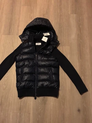 Mörkblå pufferjacka från Cevior - Snygg mörkblå pufferjacka från Cevior med avtagbar huva och dragkedja framtill. Jackan har quiltad kropp i glansigt material och stickade ärmar för en modern look. Perfekt för kyliga dagar och enkel att styla till olika outfits.