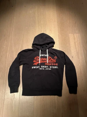 Svart Superdry hoodie  - Svart hoodie från Superdry med stor röd och vit logga samt japansk text på bröstet. Klassisk känguruficka framtill, vita dragsnören i huvan och mjukt material på insidan. Perfekt för en avslappnad streetwear-look.