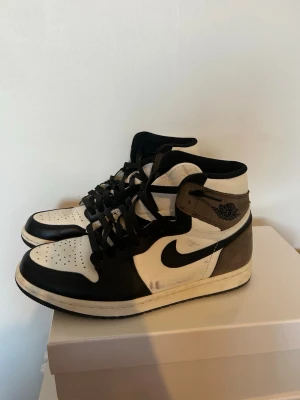 Air Jordan 1 High Mocha sneakers  - Säljer ett par Air Jordan 1 High Mocha sneakers med svart, vitt och brunt läder och mocka. Klassisk Nike Swoosh på sidan och Air Jordan-logga vid ankeln. Skorna har svarta snören och en vit mellansula. Perfekta för dig som gillar streetwear och vill sticka ut.