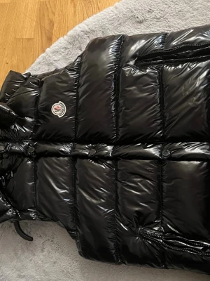 Svart glansig dunväst från Moncler - Snygg svart dunväst från Moncler med glansig finish och klassisk logotyp på bröstet. Västen har dragkedjor på fickorna och knappar framtill. Perfekt för lager-på-lager och streetstyle. Den är lite sliten men märks inte och går och fixa