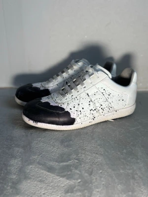 Maison Margiela svartvita sneakers - Unika sneakers från Maison Margiela i vitt läder med svarta färgstänk och svart tåparti. Klassisk låg modell med vita snören och subtila sömdetaljer. Perfekta för dig som gillar statement-plagg och konstnärlig stil.