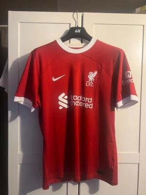 Liverpool matchtröja Alexander-Arnold - Röd Liverpool FC matchtröja direkt från shoppen utanför Anfield med vita detaljer och tryck. Framsidan har klubbmärke, Nike-logga och sponsortryck. Baksidan har namnet Alexander-Arnold och nummer 66 i vitt. Korta ärmar och rund hals. Perfekt för fotbollsfans! Köpt på plats i Liverpool utanför Anfield för 1899k