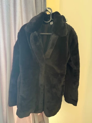 Svart fuskpälsjacka med krage - Mysig svart fuskpälsjacka med bred krage och rak passform. Jackan har en klassisk look och är tillverkad i mjukt, fluffigt material som håller dig varm under kalla dagar. Perfekt för dig som vill ha en stilren och trendig vinterjacka.