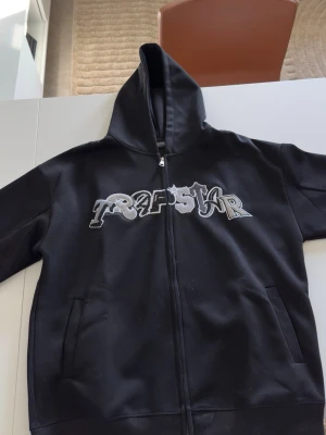  Trapstar hoodie  - Svart hoodie från Trapstar med dragkedja och broderad logga i vitt och grått framtill. Klassisk modell med huva och fickor på sidorna. Perfekt för en streetwear-look och passar till det mesta. Materialet känns mjukt och bekvämt. Den är S men lite oversized