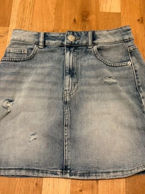 Ljusblå jeanskjol med slitningar - Trendig ljusblå jeanskjol i A-linjeform med slitna detaljer och klassiska fickor fram och bak. Kjolen har bälteshällor och knappstängning framtill. Perfekt för en avslappnad och cool stil.