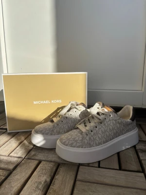 Beige sneakers från Michael Kors - Snygga beige sneakers från Michael Kors med MK-monogram över hela skon. Skorna har vita platåsulor, beige snörning och detaljer i läderimitation. Perfekta för dig som gillar stilrena och trendiga sneakers med en lyxig touch. Skorna är i storlek 37.5🩷